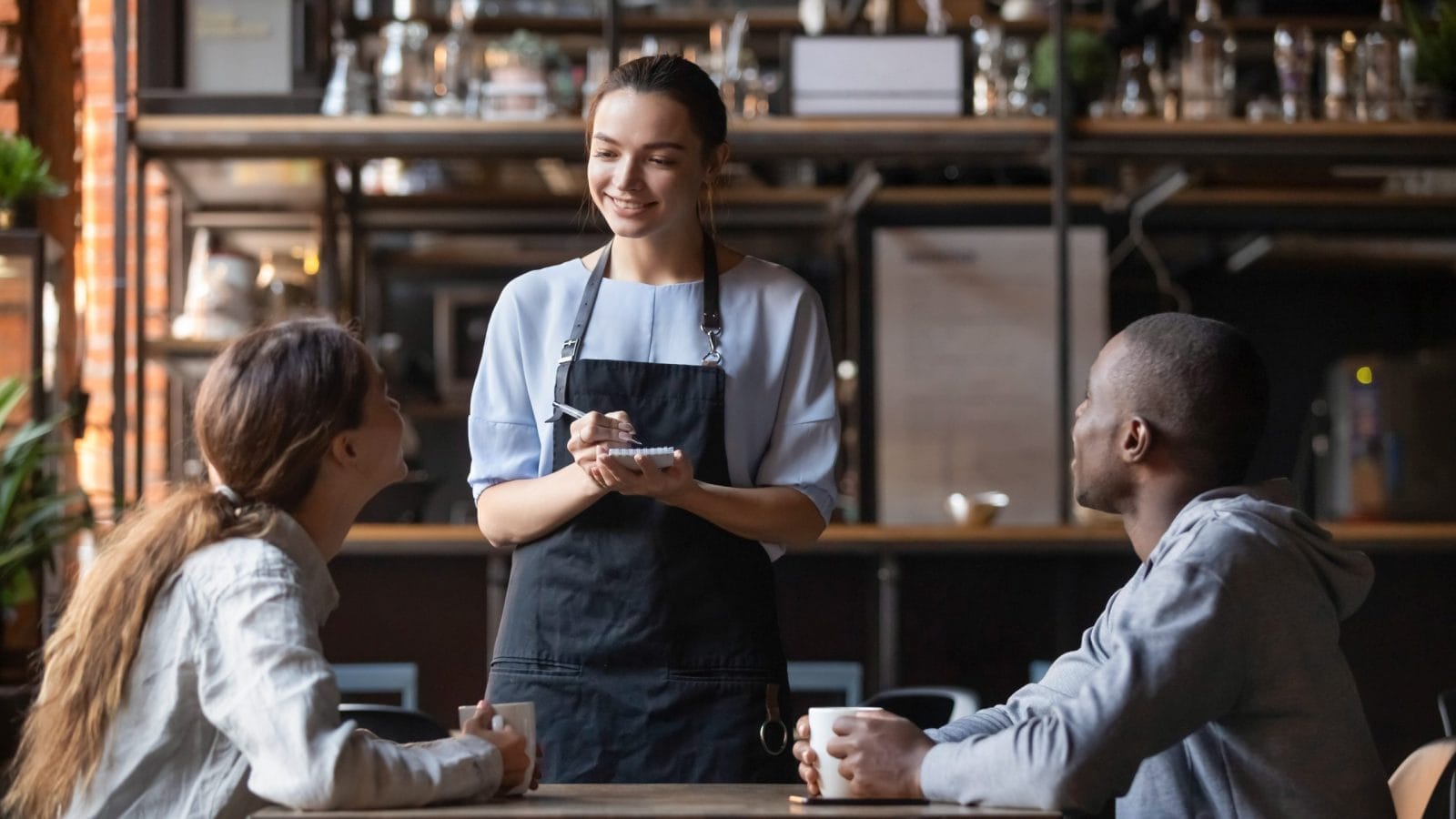 Smiling millennial waitress with notebook taking order from couple