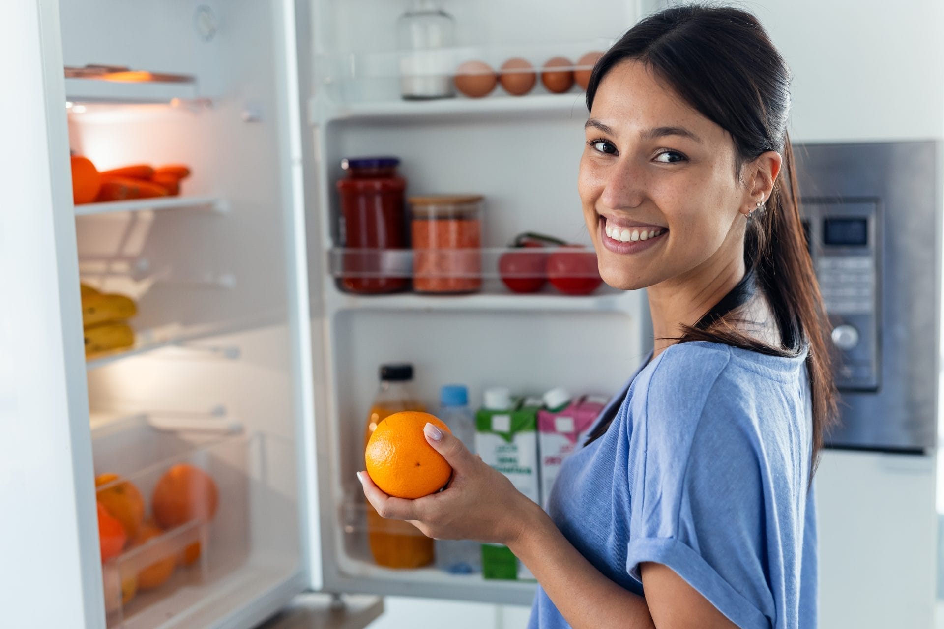 Woman standing next to open fridge holding orange