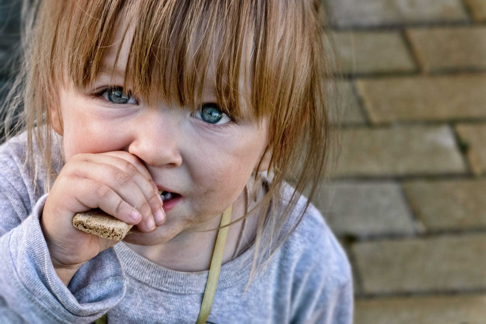 Hungry child with big clear eyes eating bread and impressively looking at the camera. Close-Up.