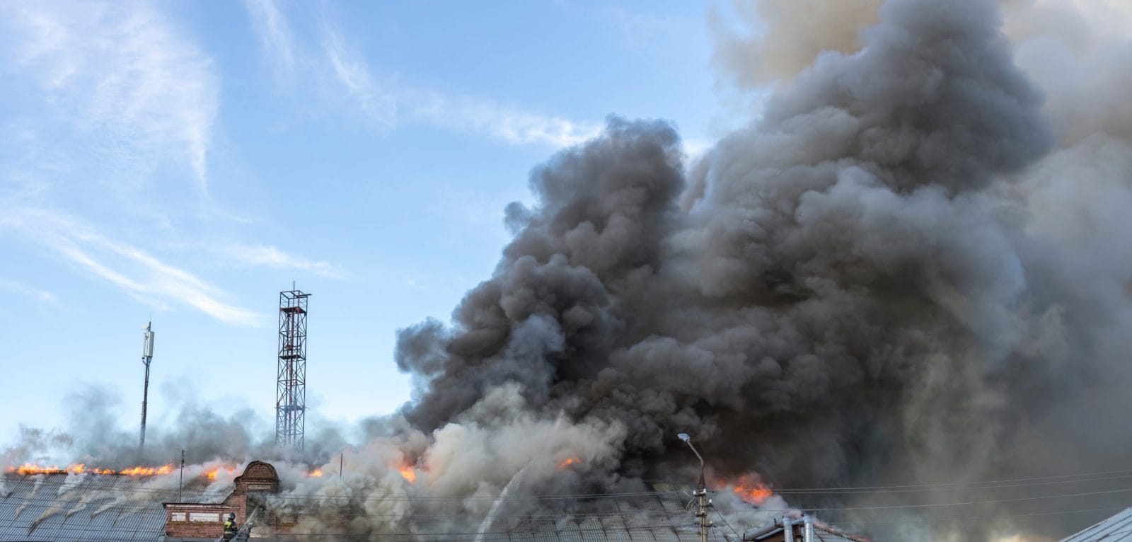 A fire is burning in the distance, with smoke billowing into the sky. The scene is chaotic and dangerous, with firefighters working to put out the flames. Scene is tense and urgent