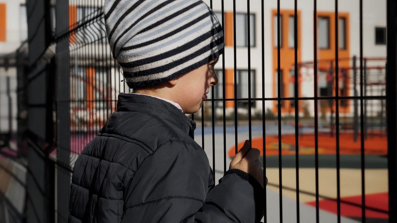 Upset lonely boy looking through metal fence on children palyground and modern school