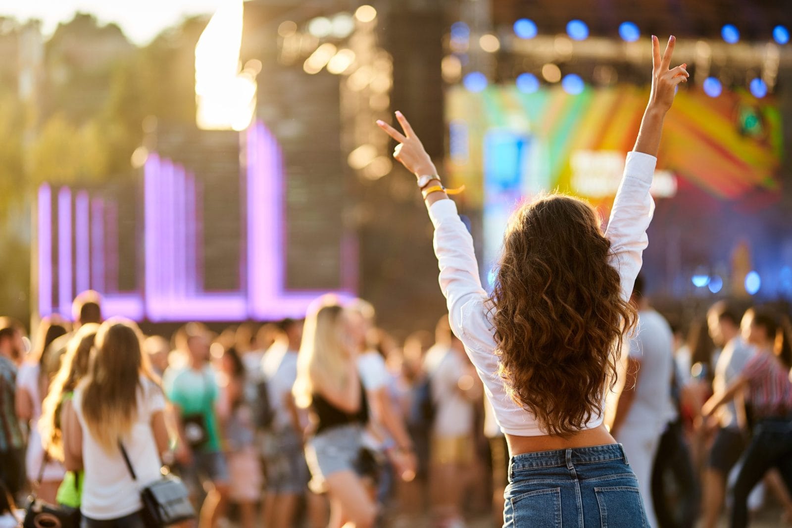 Back view of joyful woman with raised hands enjoys summer music festival. Crowd dances at beach concert, sunset light. Happy fan cheers at outdoor live event. Excited attendee celebrates, vibes.